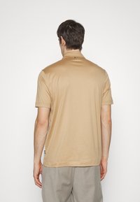 Beige kortärmad polo med en diskret krage och en liten logotyp på baksidan. Slät tyg och en avslappnad passform.