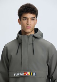 Giacca impermeabile grigia con colletto alto, cordini regolabili e una banda con logo in colori vivaci sul petto. Design moderno e fluido.