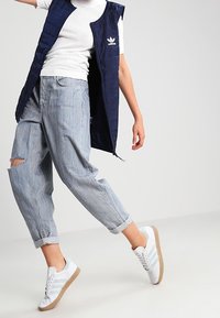 Person som bär ljusblå jeans med uppvikt ben och rivet knä, vita Adidas-sneakers med ljusgröna ränder, en vit kortärmad skjorta och en marinblå väst.