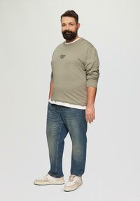 Sweat-shirt vert olive avec logo noir, superposé sur une chemise blanche, associé à un jean bleu et des baskets beiges. Look décontracté, coupe relax.