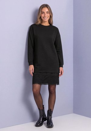 Une femme souriante porte une robe noire à manches longues avec un ourlet en dentelle, des collants noirs et des bottines devant un fond de studio lavande pâle.