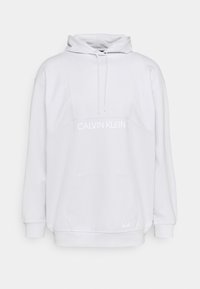 Hoodie blanc Calvin Klein avec poche avant, capuche ajustable avec cordon de serrage, et logo imprimé sur le devant.