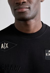 Črna majica s kapuco z vezenimi nalepkami z logotipi "A/X" in "Armani". Mehka tkanina s krojnim ovratnikom in teksturiranimi poudarki.