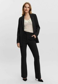 Blazer nero su una maglia leggera e texturizzata con ruffle. Pantaloni neri svasati e décolleté a punta completano l'outfit.