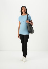 Blå ribbestrikket graviditetstopp med korte ermer, buet design ved midjen, kombinert med svarte leggings og hvite joggesko. Svart totebag holdt i den ene hånden.