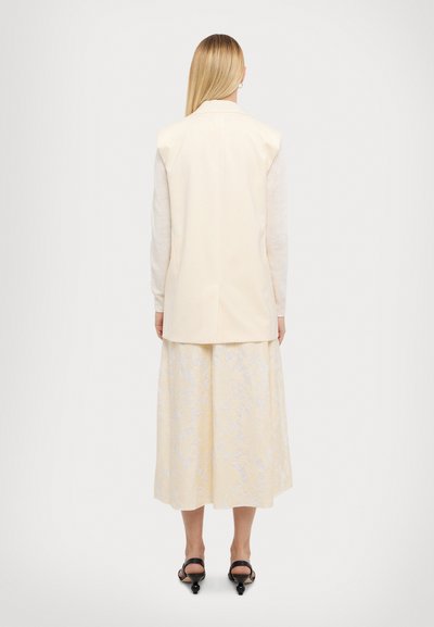 Beige ærmeløs vest med sideslids, parret med en lys gul nederdel med blomstermønster. Modellen bærer sorte højhælede sandaler.