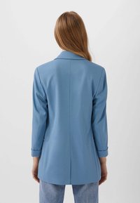Person med långt hår, klädd i en ljusblå skräddarsydd kavaj och ljusa jeans, står med ryggen mot en helt vit bakgrund.
