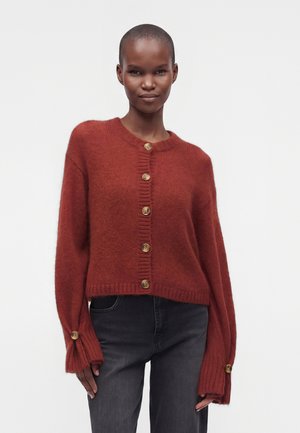 Femme portant un cardigan boutonné couleur rouille avec des manches surdimensionnées et un jean noir, debout devant un fond blanc uni.