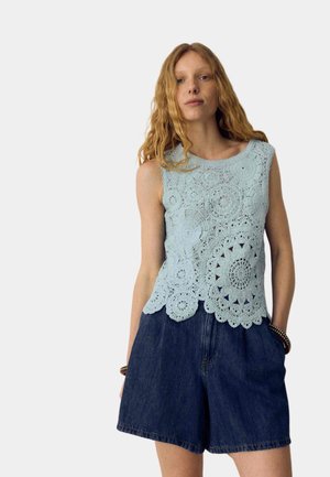 Vrouw met lang golvend haar, gekleed in een lichtblauw gehaakt mouwloos topje en donkerblauwe denim shorts, met handen in de zakken, tegen een effen lichte achtergrond.