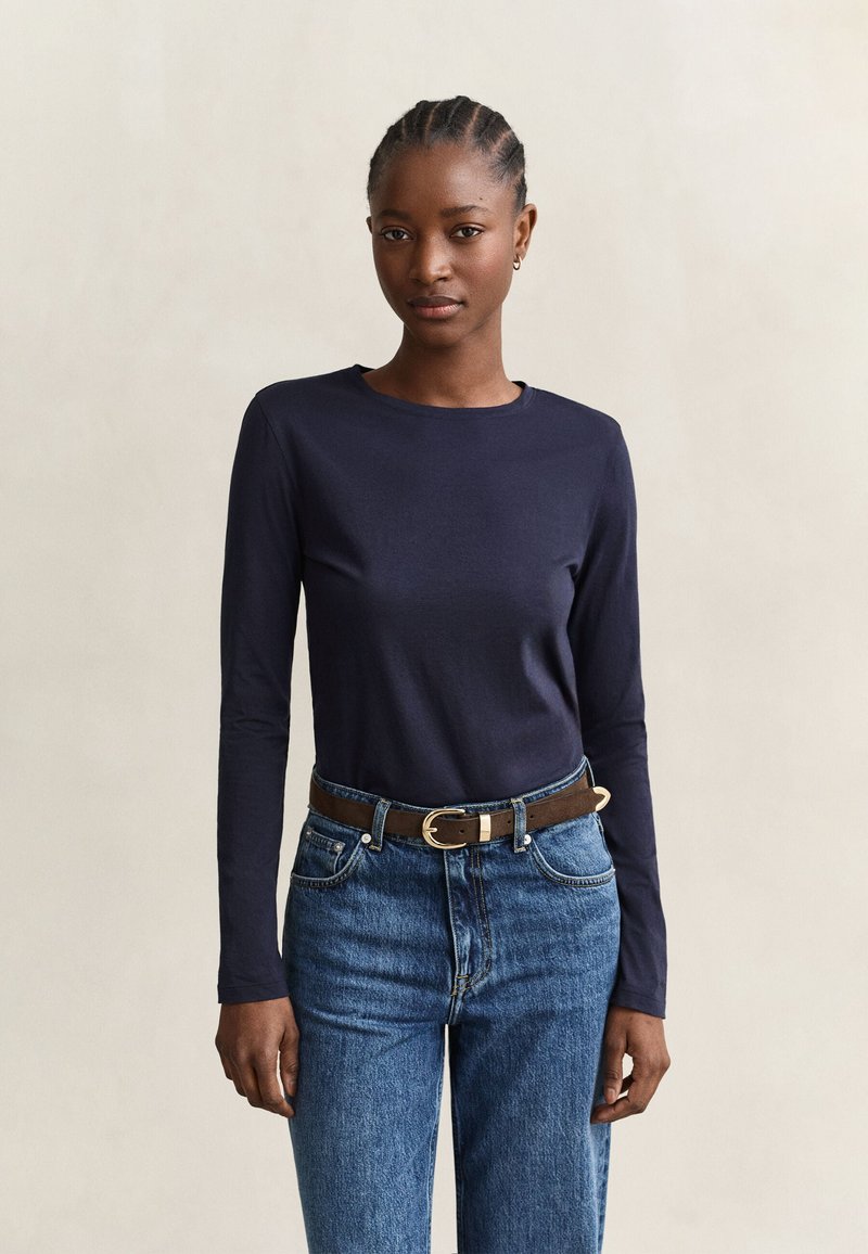 Femme aux cheveux tressés portant une chemise à manches longues foncée, un jean bleu et une ceinture marron, debout devant un fond beige uni.