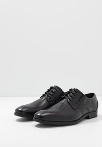Scarpe da uomo in pelle nera con finitura liscia, caratterizzate da punte tonde, design con laccetti e sottili dettagli testurizzati sui lati.