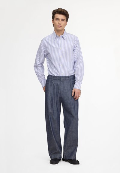Jeune homme debout portant une chemise à rayures bleues et blanches boutonnée, rentrée dans un pantalon ample bleu foncé avec des chaussures noires sur fond blanc.