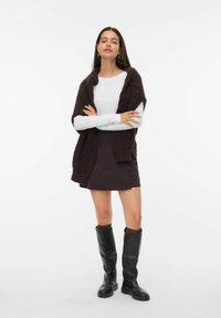 Una giovane donna sta in piedi con le braccia incrociate, indossando una camicia bianca a maniche lunghe, una mini gonna scura, stivali neri sopra il ginocchio e un maglione scuro lavorato a maglia sulle spalle.