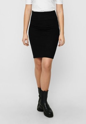 Mujer con una falda negra ajustada hasta la rodilla, top blanco de manga corta y botas negras de cuero hasta la mitad de la pantorrilla, de pie contra un fondo liso.