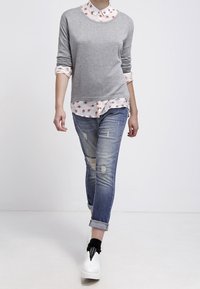 Grauer Strickpullover über einem weißen Hemd mit floralen Mustern, kombiniert mit verwaschenen blauen Jeans und weißen Sneakers mit schwarzen Akzenten.