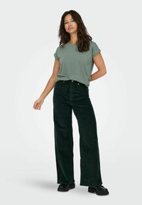 T-shirt verde a maniche corte abbinato a pantaloni larghi in corduroy verde scuro. I pantaloni presentano tasche frontali e una leggera trama a costine. Scarpe nere.