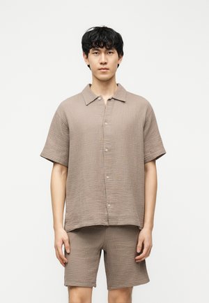 Młody mężczyzna stojący na białym tle, ubrany w luźną, teksturowaną beżową koszulę z krótkim rękawem i pasujące krótkie spodenki.