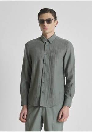 Jonge man die een groen textuur lang shirt draagt, bijpassende groene broek en donkere zonnebril, staand tegen een effen lichte achtergrond.