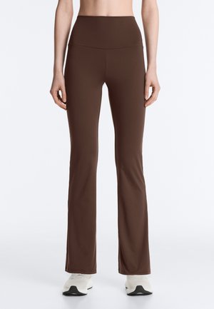 Bruine slim-fit leggings met hoge taille, gedragen met witte sportschoenen, getoond van taille tot voeten tegen een effen achtergrond.