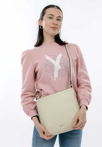 Borsa a tracolla in pelle color crema con finitura testurizzata, design piatto e tracolla regolabile, tenuta da una persona che indossa un felpa rosa.