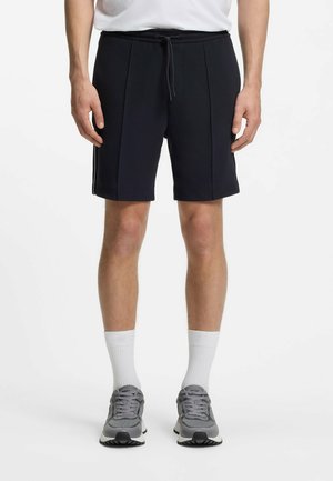 Mężczyzna w czarnych spodenkach na sznurki, białych skarpetach za kostkę, szarych butach sportowych, stojący na białym tle, ręce swobodnie opuszczone wzdłuż ciała.