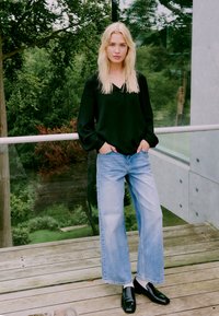 Schwarze Bluse mit Struktur und Knopfdetail, hellblaue weite Jeans und glänzende schwarze Loafers, vor einer Kulisse aus Grünanlagen und moderner Architektur.