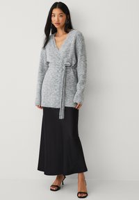 Cardigan avvolgente grigio realizzato in materiale morbido e testurizzato, con maniche lunghe e vita con lacci, abbinato a una gonna maxi nera in satin.