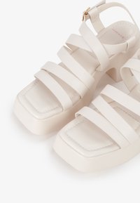 Sandali bianchi con tacco a blocco, caratterizzati da più cinturini larghi e piatti. Il design con punta quadrata presenta una soletta imbottita e una texture liscia con cuciture sottili.