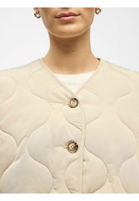 Beige gestiktejas met een geschulpte ontwerp, twee decoratieve knopen en een ronde halslijn, gemaakt van gladde stof.