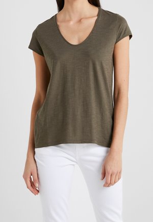 Frau trägt ein olivgrünes T-Shirt mit kurzen Ärmeln und V-Ausschnitt sowie weiße Hose, steht vor einem schlichten hellen Hintergrund.