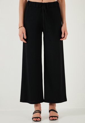 Schwarze, weite Hose aus strukturiertem Stoff mit elastischem Bund und Kordelzug. Kombiniert mit schwarzen, schmalen Sandalen.