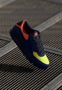 Nike Air Max-sko med en marinblå överdel, ljusa orange och gula accenter, mesh och syntetiska material, samt en synlig luftfjädring i sulan.
