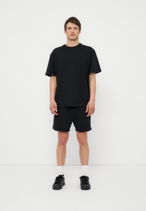 Mann steht vor einer weißen Wand, trägt ein schwarzes oversized T-Shirt, schwarze Shorts, weiße Socken und schwarze Turnschuhe, schaut nach vorne und hat die Arme entspannt hängen.