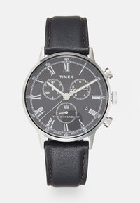 Montre avec un bracelet en cuir noir et un boîtier en acier inoxydable argenté. Le cadran noir présente des chiffres romains en argent, des sous-cadrans et un affichage de la date.