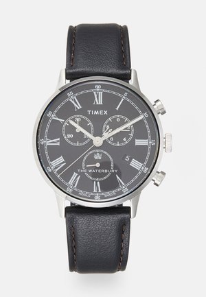 Montre avec un bracelet en cuir noir et un boîtier en acier inoxydable argenté. Le cadran noir présente des chiffres romains en argent, des sous-cadrans et un affichage de la date.