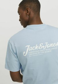 Camiseta de algodón azul claro con mangas cortas. La parte trasera presenta texto en blanco: "Jack & Jones Essentials Dept. Worldwide" y otros detalles.