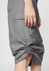 Lys grå cargo shorts med snøring i kanten, som har en sidelomme og sammenkrøpet stoffdetalj nær kantene. Glatt tekstur.