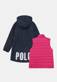 Marinblå huvad jacka med "POLO" i stora vita bokstäver på ryggen, parat med en rosa vadderad ärmlös väst med ett kviltat mönster.