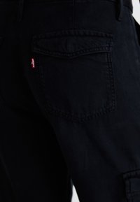 Crne traperice od denima s dvostrukim stražnjim džepovima i crvenom Levi's oznakom. Imaju klasičan kroj i čvrstu teksturu tkanine.