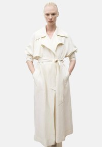 Vit linne trench-coat med bälte i midjan, överdimensionerade ärmar, stor krage och djupa sidofickor. Slät textur, minimalistisk design.
