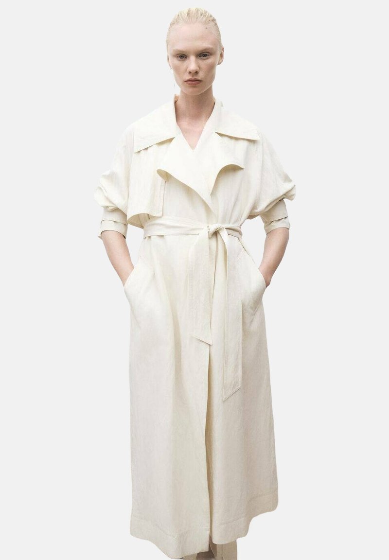 Vit linne trench-coat med bälte i midjan, överdimensionerade ärmar, stor krage och djupa sidofickor. Slät textur, minimalistisk design.