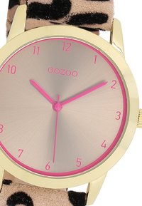 Montre dorée avec un cadran rose métallique, des index rosés et des aiguilles. Le bracelet présente un motif léopard beige pour ajouter de la texture.