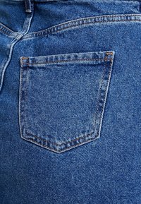 Snímek blízkého pohledu na modré džíny z denimu, které mají zadní kapsu s kontrastním oranžovým prošíváním, texturovanou tkaninu a mírně vybledlý povrch.
