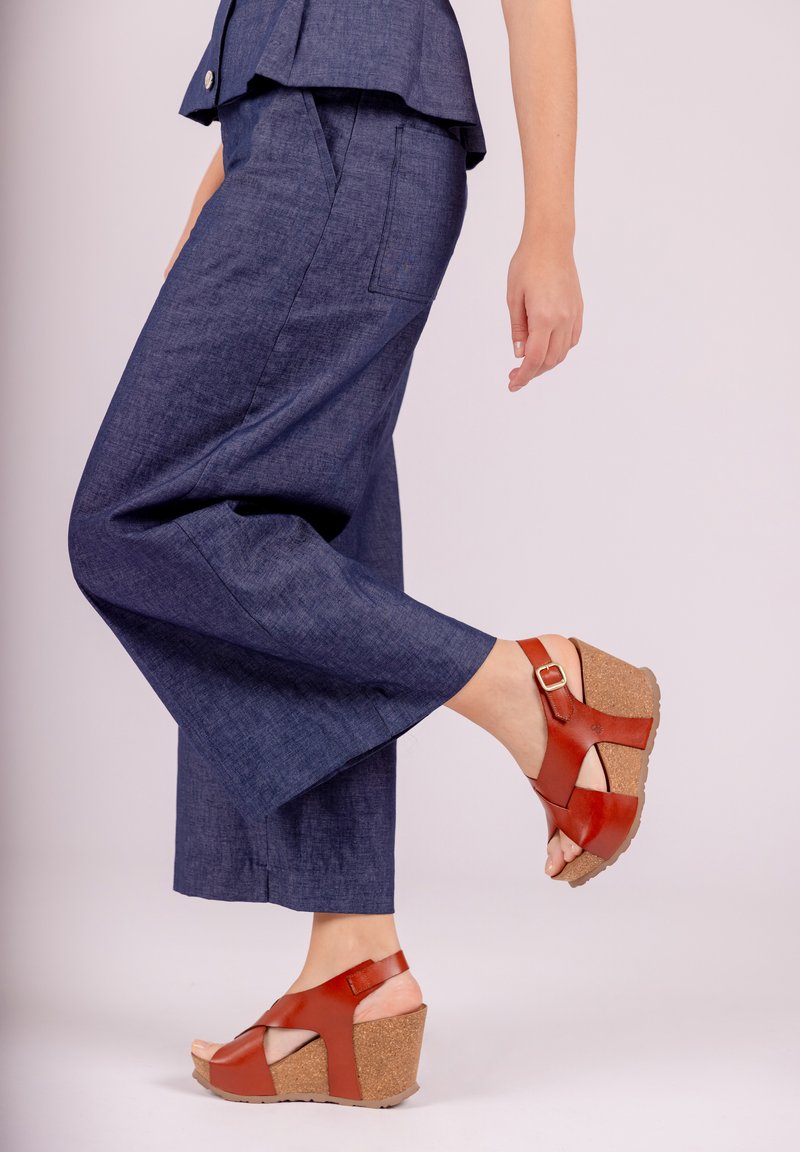Brown wedge sandals with cork soles and cross-strap design; worn with dark blue, loose-fitting cropped pants. Smooth fabric texture.