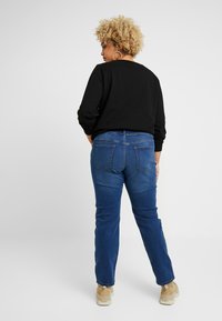 Camisa negra de manga larga, jeans rectos azules con bolsillos traseros y zapatillas beige, vistos desde la parte posterior contra un fondo neutro.