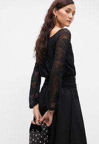 Μαύρη δαντελένια μπλούζα με μακριά μανίκια, λουλουδάτο σχέδιο και άνετη εφαρμογή. Συνδυασμένη με μαύρη φούστα και μαύρη τσάντα με καρφιά.