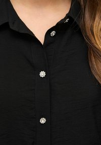 Crna košulja na dugmad s teksturiranim materijalom, klasičnom kragnom i ukrasnim cvjetnim gumbima sa prozirnim draguljima.