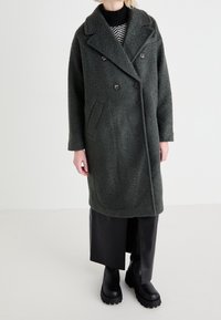 Abrigo de lana de doble faz en verde oscuro con cuello con muesca, bolsillo lateral y acabado texturizado, combinado con pantalones de cuero negros y botas.