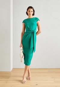 Zelené šaty do délky kolen s detailem na zavázání v pase, krátké rukávy typu cap sleeve, vyrobené z hladkého materiálu. Modelka drží bílou kabelku a má na sobě sandály na pásky.