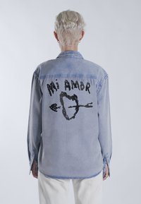Világoskék farmering hosszú ujjakkal, fekete grafikával egy szívről és nyilról, felette "Mi Amor" felirattal.